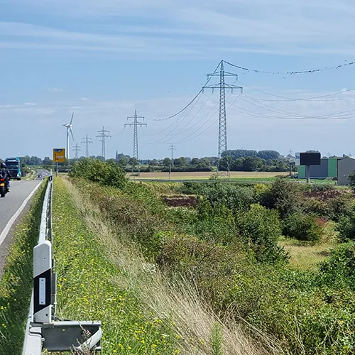 Straße mit Lkw, rechts Böschung und Strommasten, Windräder in der Ferne