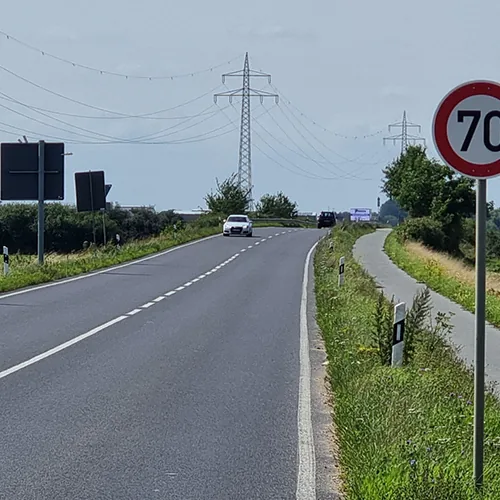 Landstraße mit Tempo-70-Schild und Radweg, Strommasten und Bäume am Horizont