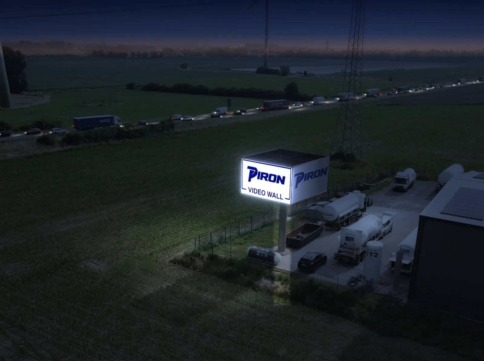 Dieselbe Ansicht in der Dämmerung: die LED-Fläche der Wall leuchtet hell über Feld und Hof, auf dem Tweestrom fahren Lkw und Pkw mit eingeschaltetem Licht
